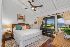 A bedroom with a bed, bedside lamps, a ceiling fan, a view of the outdoors, and a wicker table with a plant.