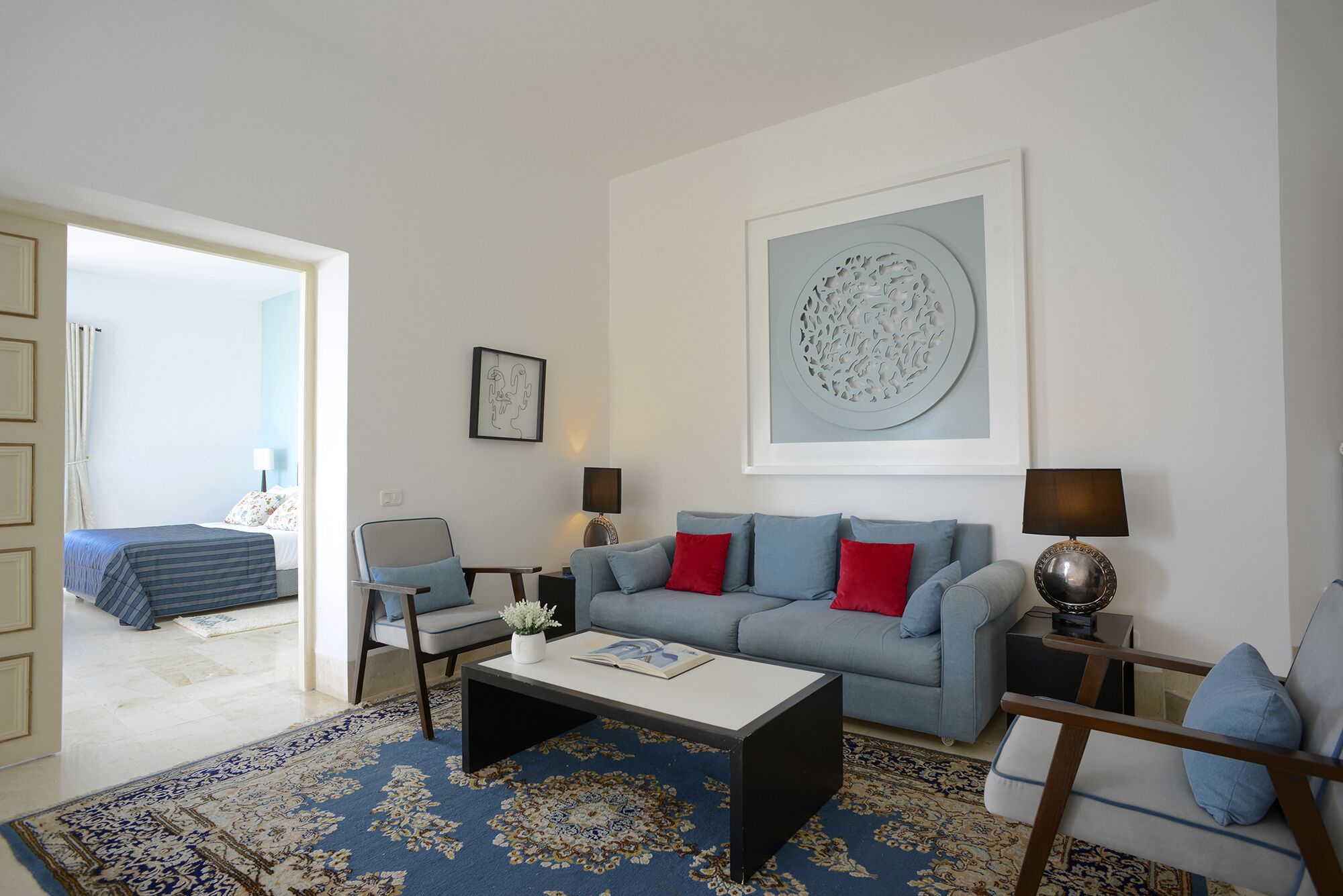 Een woonkamer met een bank, salontafel en vloerkleed. Een deur leidt naar een andere kamer.
