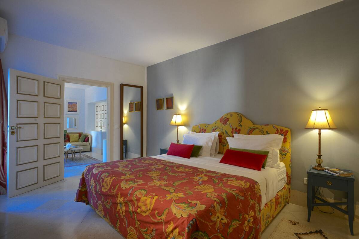 Quarto com uma cama grande, duas luminárias de cabeceira, mesa de cabeceira e uma porta que dá para outro espaço.