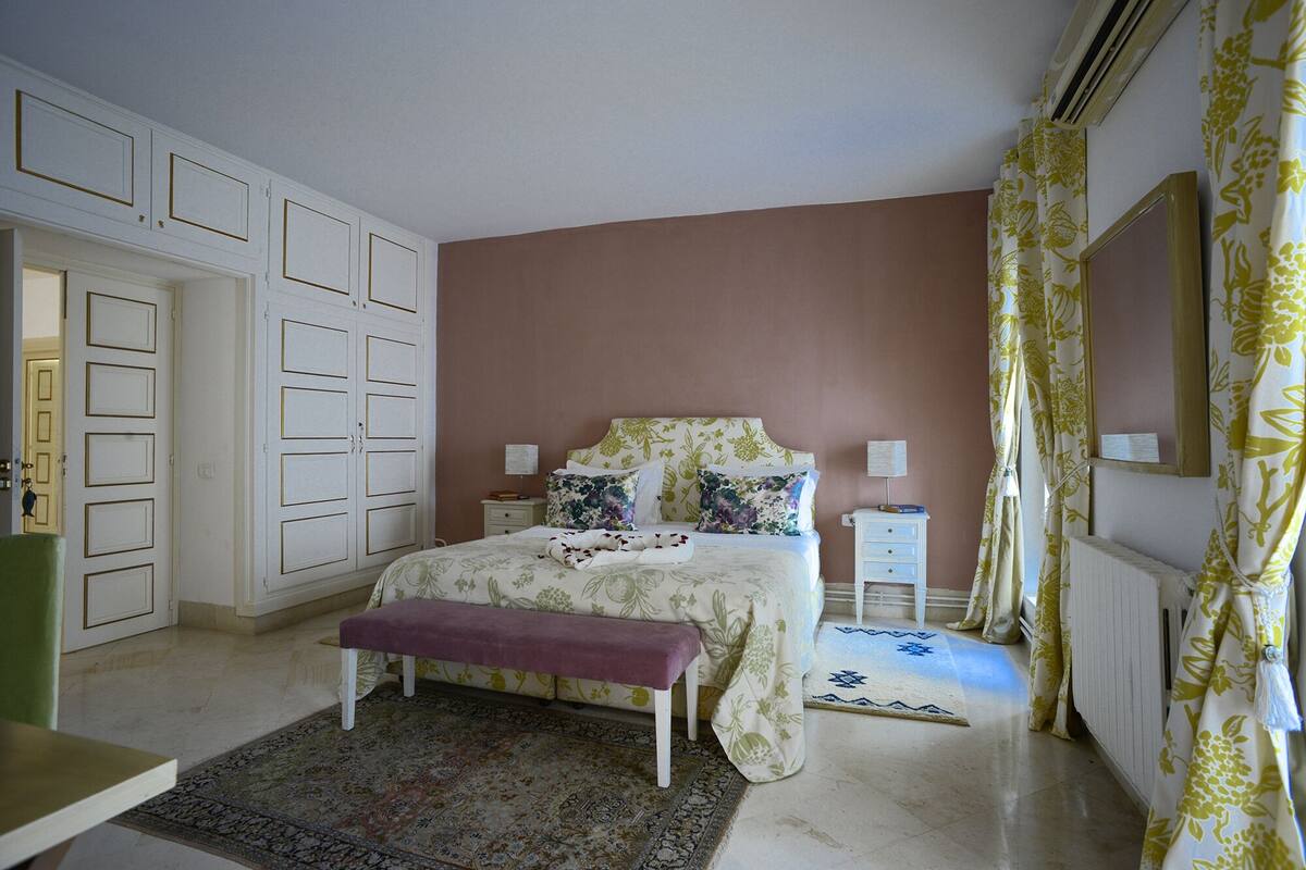Quarto com cama, um banquinho, mesa de cabeceira e cômoda.