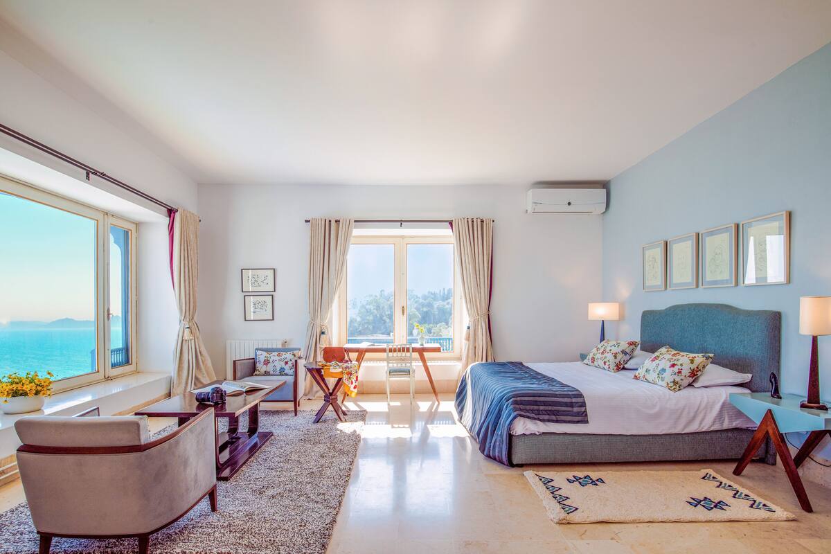 Quarto com cama, escrivaninha, cadeira, vista para o mar e janela com cortinas.