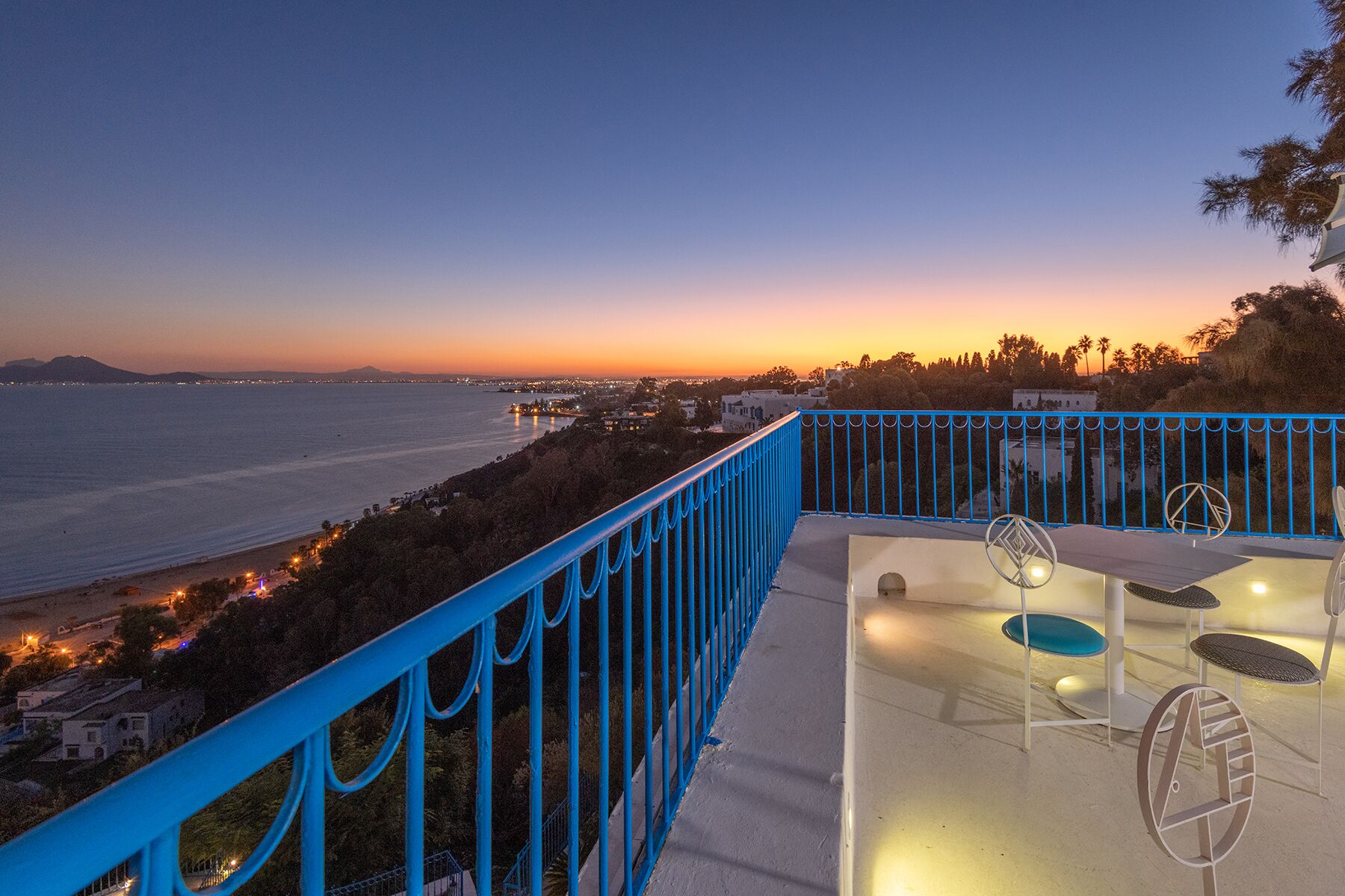 Un balcon avec vue sur l’océan au coucher du soleil, une rambarde bleue et du mobilier d’extérieur.