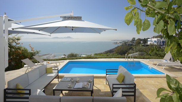 A poolside area with lounge chairs and umbrellas, a view of the sea, and a mountainous landscape in the background.