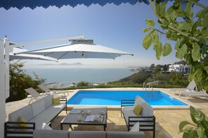 Zona junto a la piscina con sillones y sombrillas, vista al mar y paisaje montañoso al fondo.