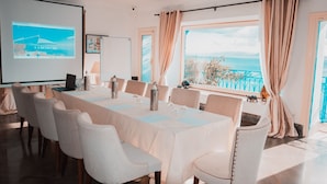 A conference room with a long table, chairs, a projector screen, and a view of the sea through large windows.