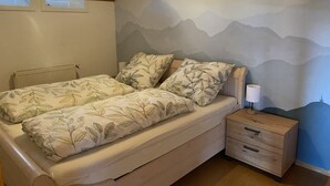 Un dormitorio con cama, mesita de noche, lámpara, radiador y un mural de montañas en la pared.