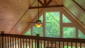 Um quarto com teto inclinado, vigas de madeira, uma janela com vista para vegetação e um lustre.