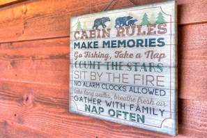 A wooden cabin with a sign listing cabin rules such as 'Go Fishing, Take a Nap, Count the Stars, Sit by the Fire, No Alarm Clocks Allowed, Take Long Walks, Breathe Fresh Air, Gather with Family, Nap Often'.