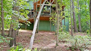 A cabin with a wooden deck and a green exterior, surrounded by trees and a forest floor.