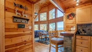 L’intérieur d’une cabane avec des murs en bois, un coin repas comprenant des chaises et une table, une cheminée et une vue dégagée sur l’extérieur grâce à de grandes fenêtres.