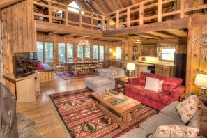 Eine geräumige Hütte mit hoher Decke, Holzvertäfelung, einem Essbereich mit Stühlen, einer Couch, einem Fernseher und einer Küchenzeile.