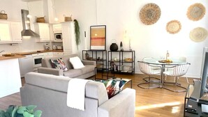 A modern living room with a grey sofa, a coffee table, and a dining area with a glass table and metal chairs. There is a kitchen area with white cabinets and a microwave. - (008) Cosy & Chic Old Town (Antibes)