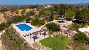 A large swimming pool with a sun terrace and a covered seating area surrounded by trees and a hilly landscape. - Quinta Molinum Ad Mare | 8 Bedrooms | Sea Views | Boliqueime (Boliqueime)