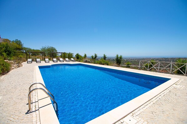 Una piscina rectangular con agua azul cristalina, rodeada por una terraza de piedra y una pérgola con sillones de descanso.