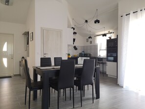 Uma área de refeições moderna com uma mesa retangular e seis cadeiras, uma cozinha com forno embutido e micro-ondas, e um lustre.