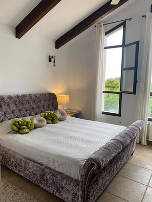 Une chambre Ă coucher avec un grand lit, une table de chevet, une lampe et une fenĂȘtre avec des rideaux.