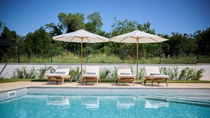 A poolside area with four lounge chairs and two umbrellas, surrounded by greenery and a clear sky.