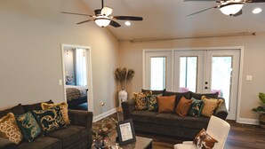 A living room with two sofas, a coffee table, and a white armchair. There are ceiling fans, a vase with dried plants, and a welcome sign on the coffee table.
