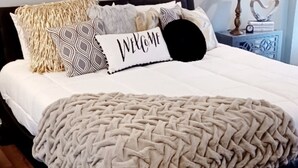 A bed with a beige blanket, multiple patterned pillows, and a 'WELCOME' pillow.