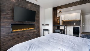 A modern bedroom with a fireplace, a TV mounted on the wall, and a kitchenette in the adjacent space.