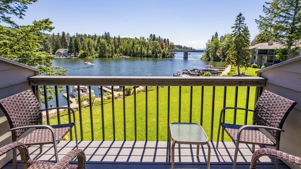 A balcony with wicker chairs, a small table, and a view of a lake with boats, a bridge, and forested land.