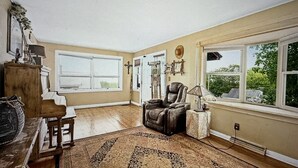 A spacious living room with a large window, a leather armchair, a wooden cabinet, and a patterned rug.