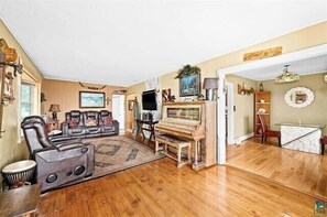A spacious living room with a wooden floor, a piano, a sofa set, and a television.