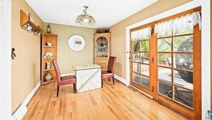 A dining area with a wooden table and chairs, a chandelier, and a large window with lace curtains.