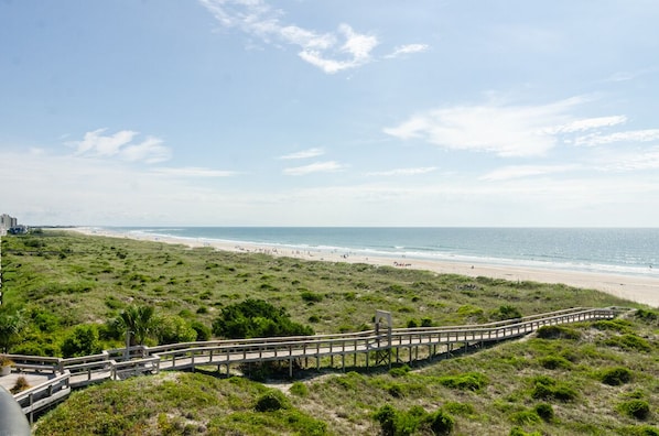 A beach with a wooden boardwalk leading to the shore, grassy dunes, and a clear sky.