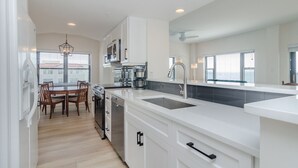 A modern kitchen with white cabinetry, a stainless steel dishwasher, and a dining area with wooden chairs. - Stunning oceanfront end unit in Duneridge! (Wrightsville Beach)