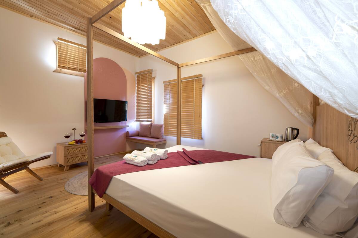 Una camera da letto con soffitto in legno, un letto con lenzuola bianche, un comodino in legno, una sedia e una finestra con persiane in legno.