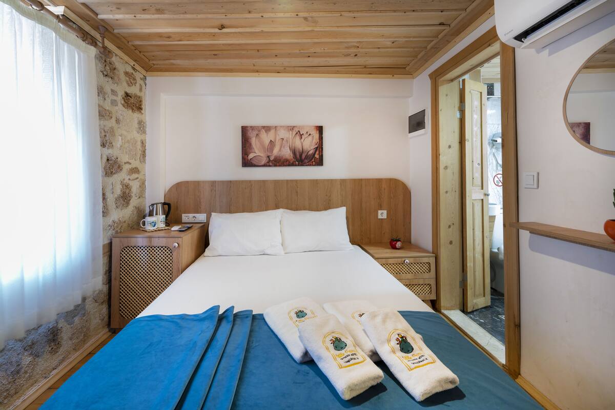 Camera d'albergo con soffitto in legno, un letto con lenzuola bianche, una coperta blu, due asciugamani con un logo, un quadro appeso alla parete e un comodino in legno con una caffettiera.