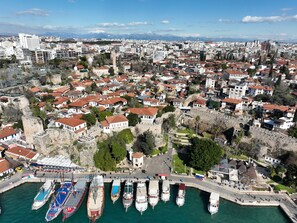 Vista aérea de un pueblo costero con edificios históricos, un puerto deportivo con barcos y un telón de fondo montañoso.