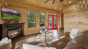 A cozy cabin interior with a fireplace, leather sofa, and a television displaying a scenic landscape. - 4King Suites | Hot Tub | Game Room | Sunrise Views (Gatlinburg)