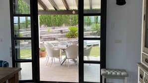 Une pièce dotée d’une porte-fenêtre coulissante donnant sur une terrasse extérieure avec une table et des chaises, un banc en bois et un téléviseur fixé au mur.