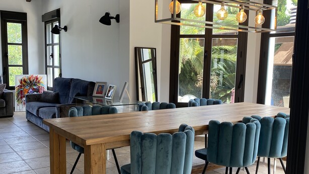 Un vaste espace dédié à la salle à manger, avec une longue table en bois et des chaises en velours bleu, de grandes fenêtres laissant entrer la lumière naturelle et un design moderne au niveau du plafond.