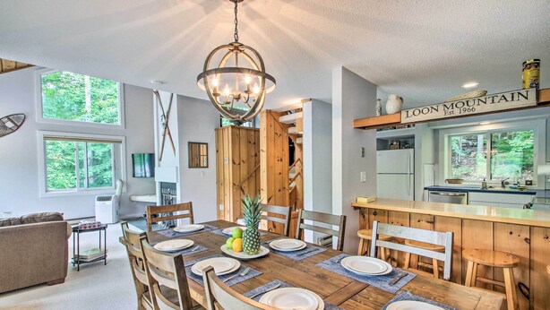 A dining area with a wooden table set for six, a chandelier, and a kitchen visible in the background. - Cozy Lincoln Condo ~ 1 Mi to Loon Mountain! (Lincoln)