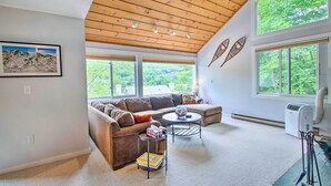 A spacious living room with a high ceiling, large windows offering a view of greenery, a brown sofa, a small coffee table, and a standing heater. - Cozy Lincoln Condo ~ 1 Mi to Loon Mountain! (Lincoln)