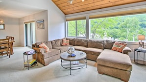 A spacious living room with a large sectional sofa, a coffee table, and a view of a forest through large windows. - Cozy Lincoln Condo ~ 1 Mi to Loon Mountain! (Lincoln)