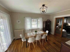 Ein Essbereich mit Holztisch, weißen Stühlen, einem Heizkörper, einem Fenster mit Vorhängen und einer Deckenleuchte.