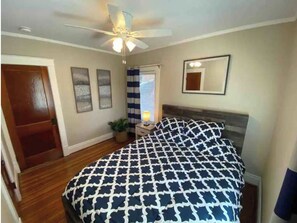 Ein Schlafzimmer mit Bett, Deckenventilator, Fenster mit Vorhängen, Nachttisch mit Lampe und Spiegel an der Wand.