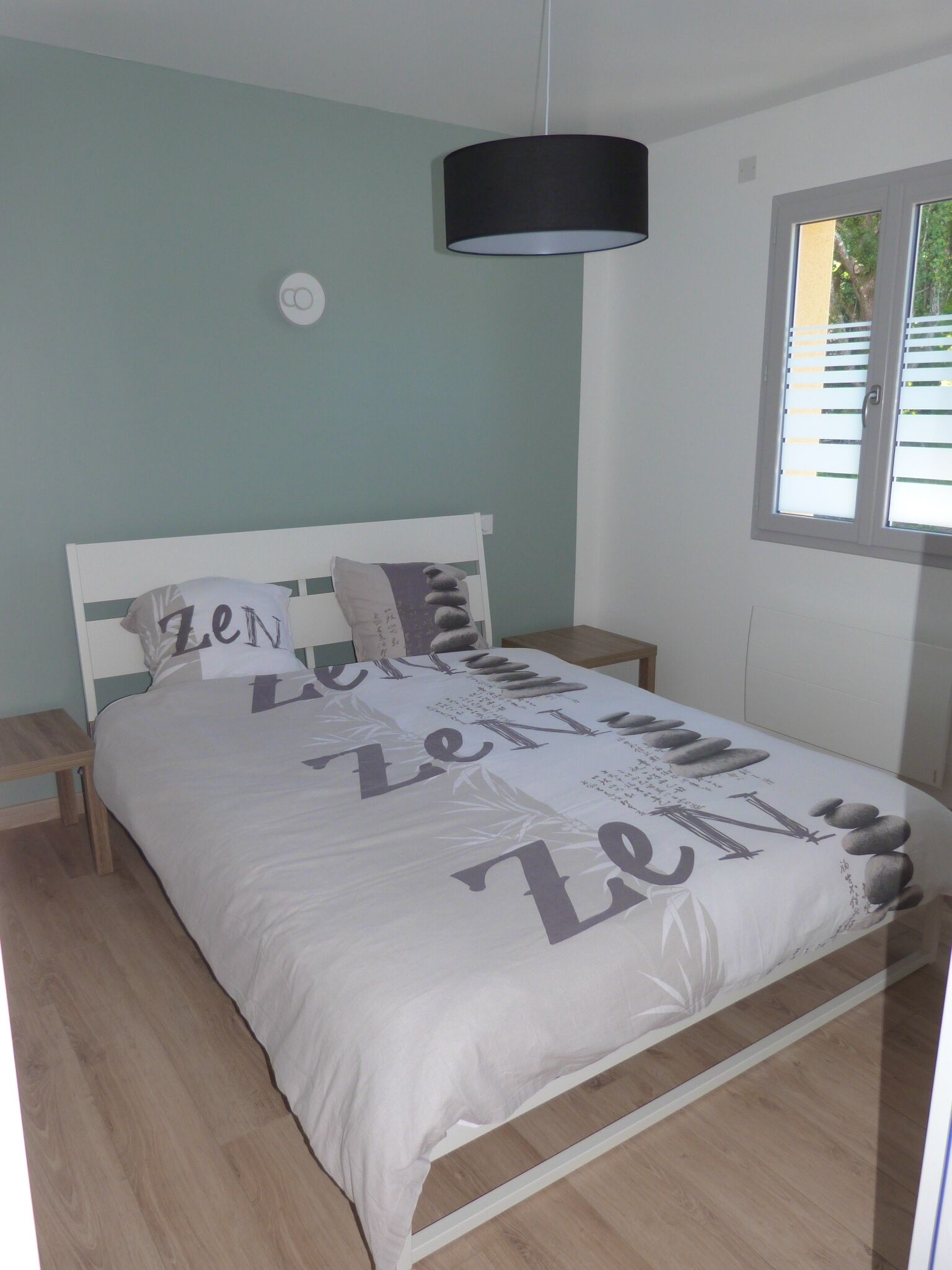 A bed with a white headboard and a bedspread with Zen text, a black hanging lamp, and a window with blinds.