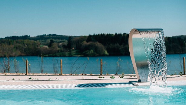 A swimming pool with a water feature and a view of a lake and forest in the background.