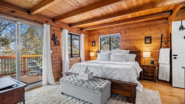 A wooden cabin bedroom with a large bed, a bench, a desk, and a balcony with chairs. - Spacious, Modern Mountain Cabin with Hot Tub (Running Springs)