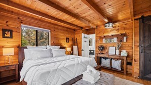 A wooden cabin bedroom with a large bed, bedside lamps, a bench, and a view of the outdoors. - Spacious, Modern Mountain Cabin with Hot Tub (Running Springs)