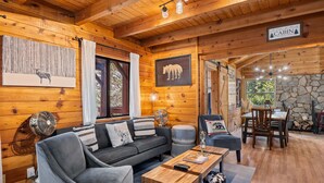 A cabin interior with a wooden interior, a sofa, a dining table, and a stone fireplace. - Spacious, Modern Mountain Cabin with Hot Tub (Running Springs)