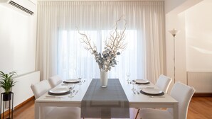Uma sala de jantar com uma mesa branca posta para dois, um vaso com galhos e um abajur.
