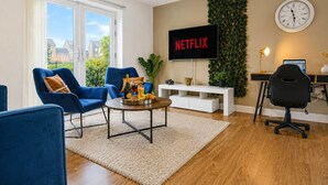 A modern living room with a flat-screen TV, blue chairs, a coffee table with fruit, and a desk with a computer.