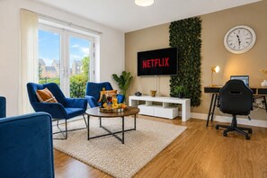 A modern living room with a flat-screen TV, blue chairs, a coffee table with fruit, and a desk with a computer.
