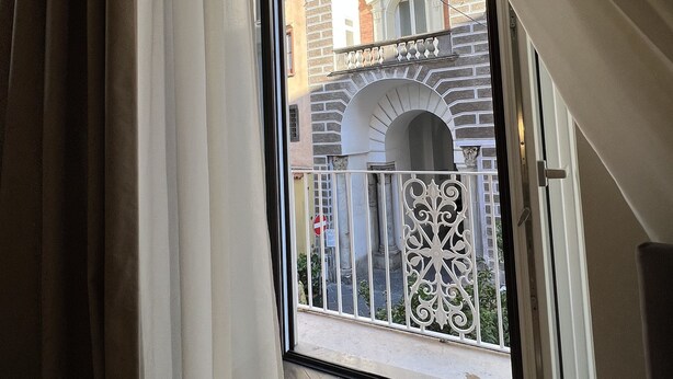 A view through a window with curtains, revealing a balcony with a metal railing and a brick building with arches in the background. - Appartamento nel Centro Storico di Sorrento, Nuovo, con Vista (Sorrento)